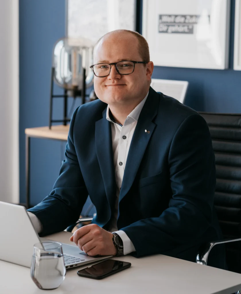 Johannes Schlemmer, Finanzexperte für Familien-Versicherung, Finanzberatung und Investitionsplanung, sitzt im Büro an einem Laptop und lächelt in die Kamera.